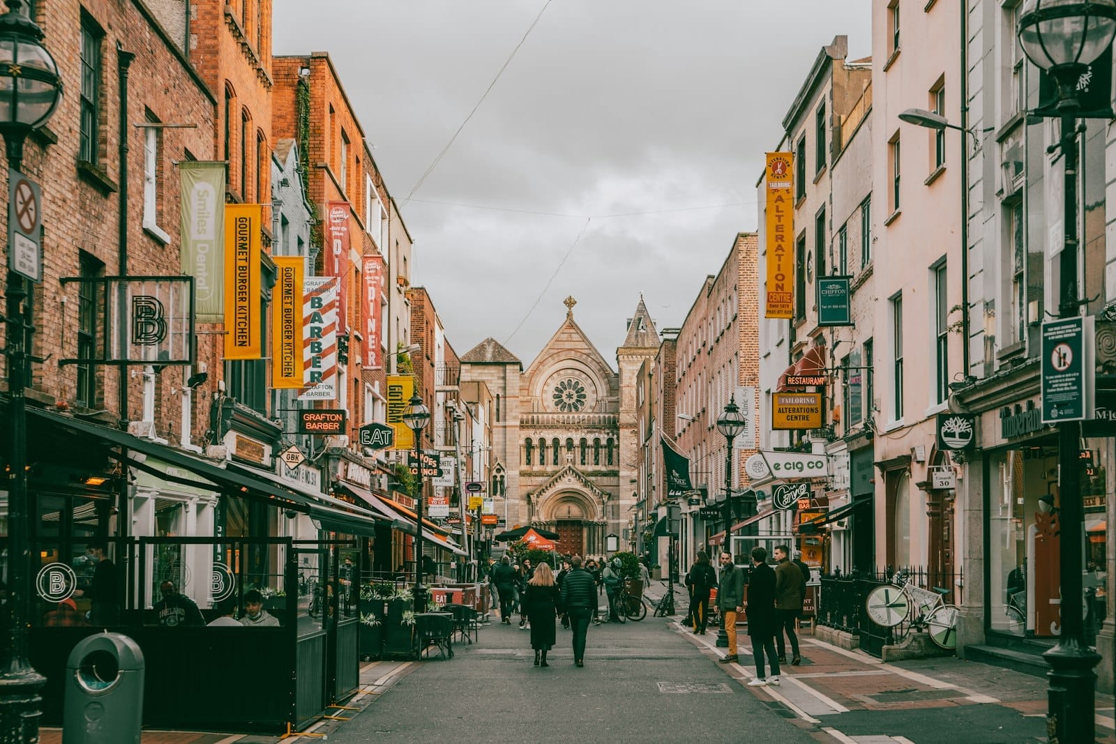 Cena de rua com pedestres em Dublin, pessoas caminhando em frente a cafés e lojas, fachadas coloridas e uma igreja histórica ao fundo.