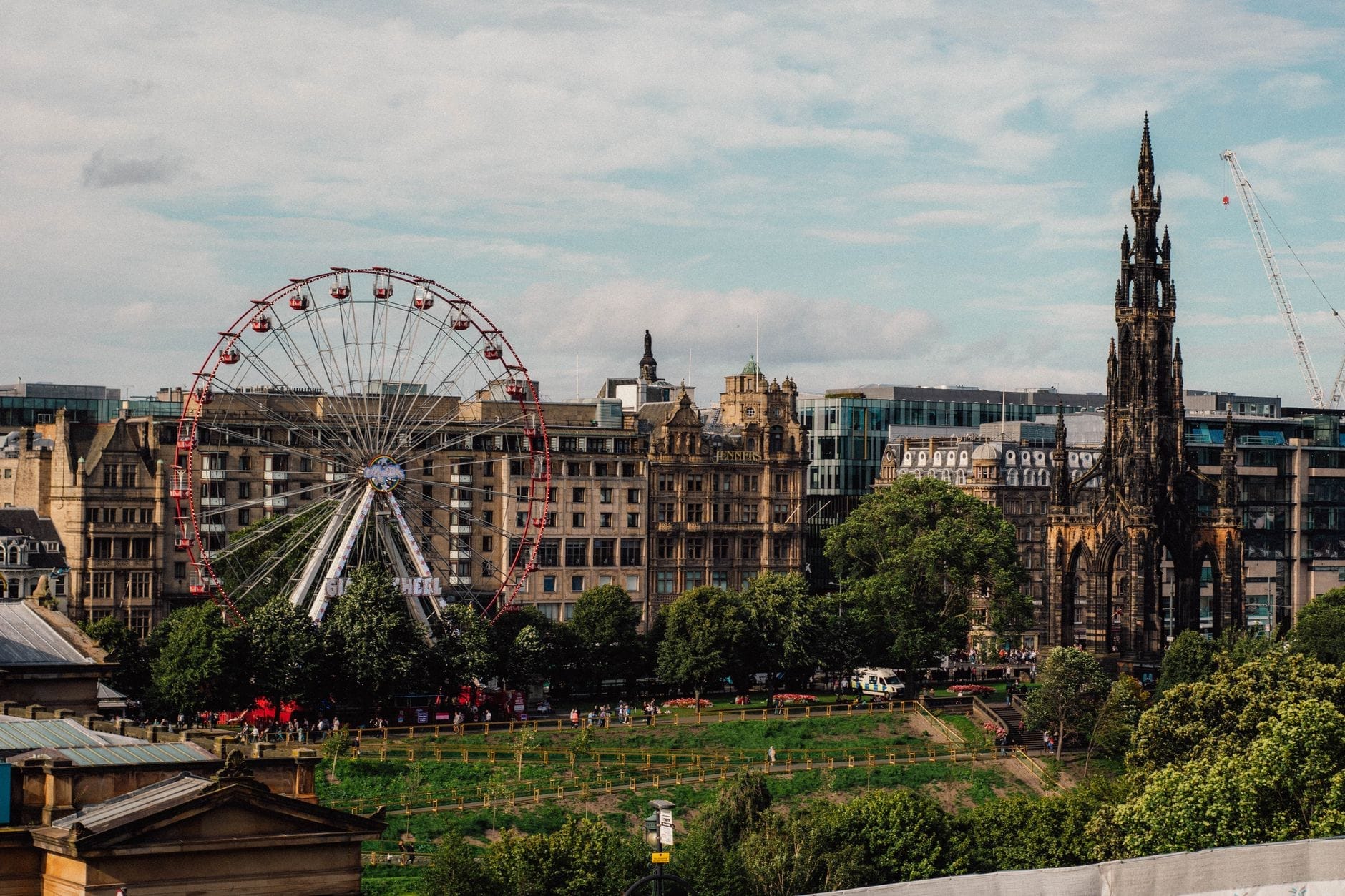Blick auf die Skyline von Edinburgh mit dem Scott Monument, einem Riesenrad, einem üppigen grünen Park und historischen Steingebäuden unter einem teils bewölkten Himmel.