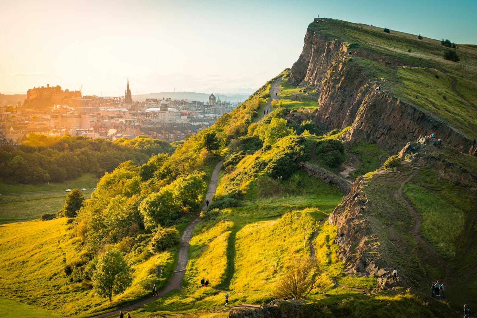 Sonnenbeschienene Aussicht auf Arthur's Seat mit der Stadt Edinburgh und dem Schloss im Hintergrund, grüne Pfade winden sich den Hang hinauf.