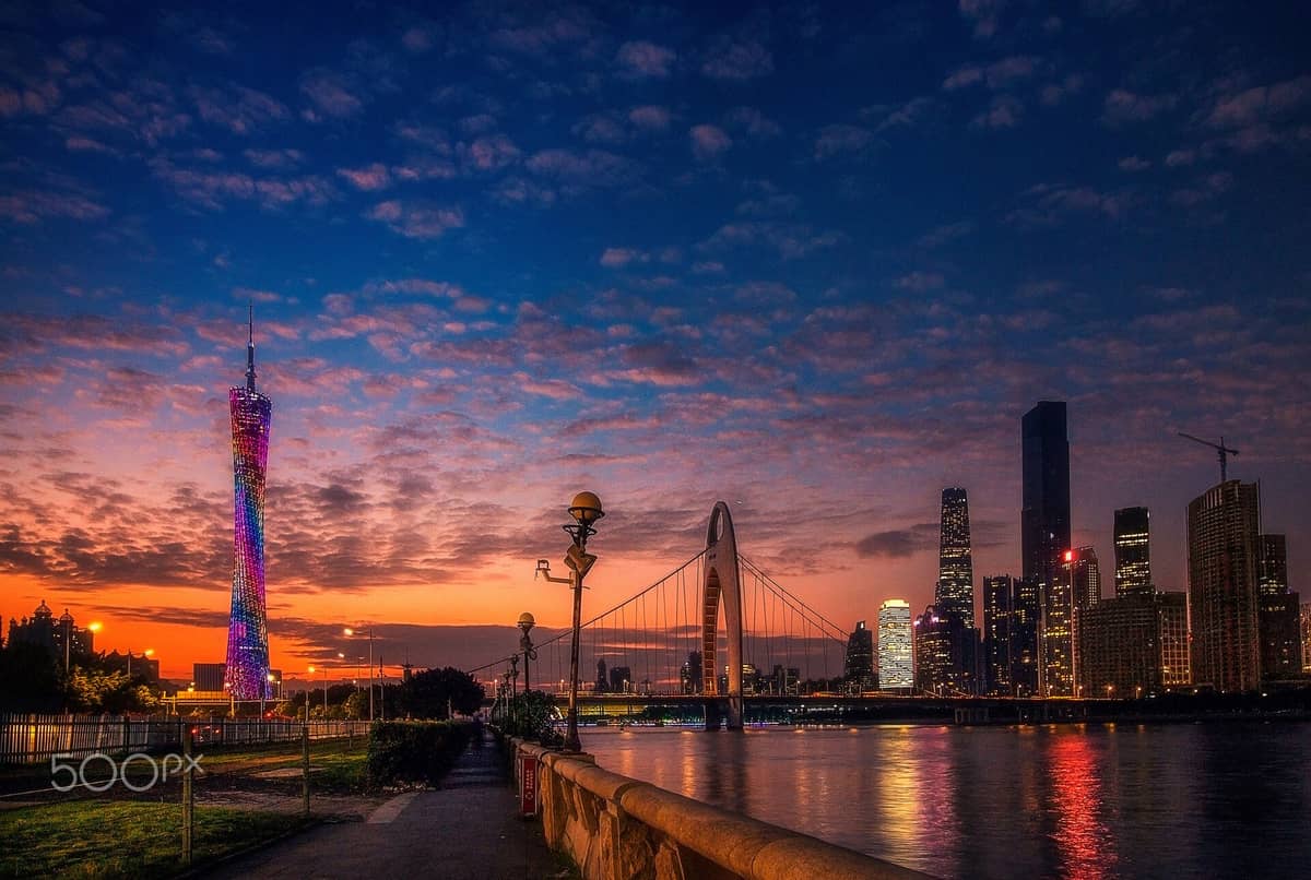 Night view of Guangzhou’s Zhujiang New Town skyline with Canton Tower illuminated, Liede Bridge, and riverside promenade under a vivid sunset sky.