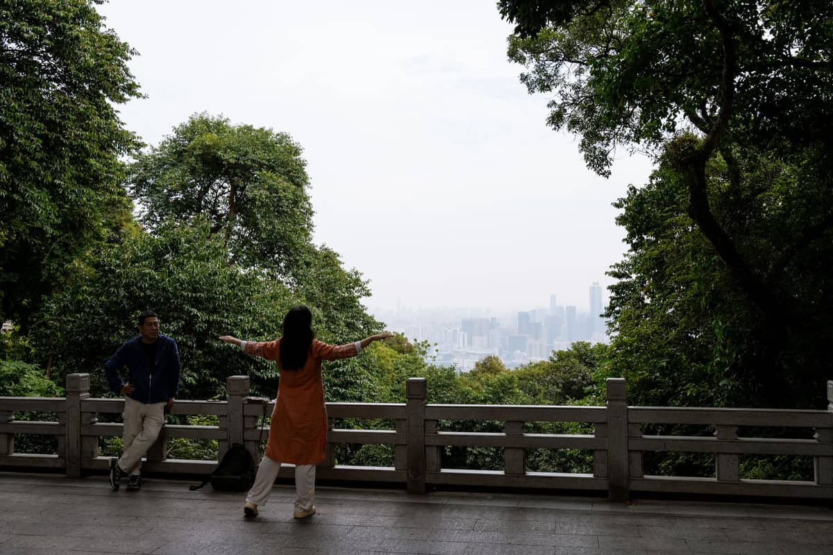 Two people stand on a stone terrace overlooking lush trees and a hazy cityscape, suggesting a scenic Guangzhou viewpoint.