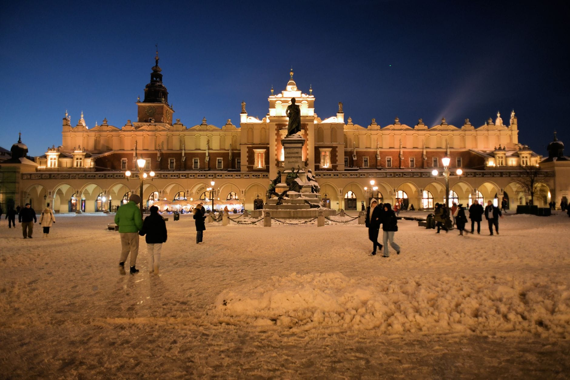 จัตุรัสหลัก Rynek Główny ในเมือง Kraków ยามค่ำคืนท่ามกลางหิมะ ผู้คนเดินผ่านไปมา และอาคาร อาคาร Sukiennice ที่สว่างไสวพร้อมอนุสาวรีย์ Adam Mickiewicz