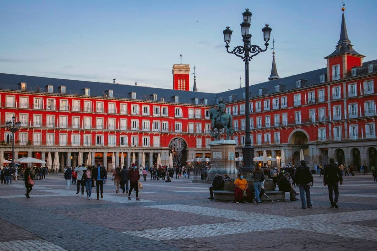 Wieczorny widok na Plaza Mayor w Madrycie z czerwonymi budynkami, centralnym pomnikiem konnym, latarnią oraz ludźmi spacerującymi i siedzącymi na placu.