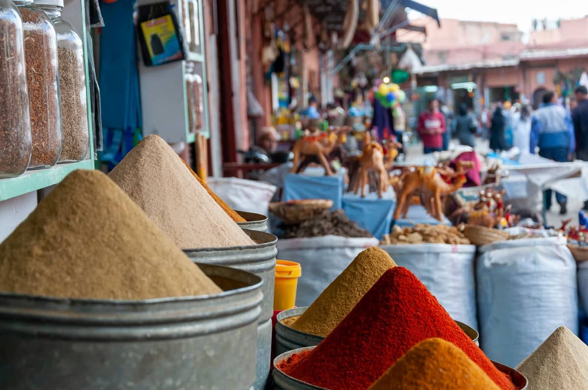 กรวยเครื่องเทศหลากสีสันในภาชนะโลหะขนาดใหญ่ที่วางแสดงอยู่ในตลาด Marrakech ที่คึกคัก โดยมีนักช้อปและสินค้าต่างๆ ให้เห็นในพื้นหลัง