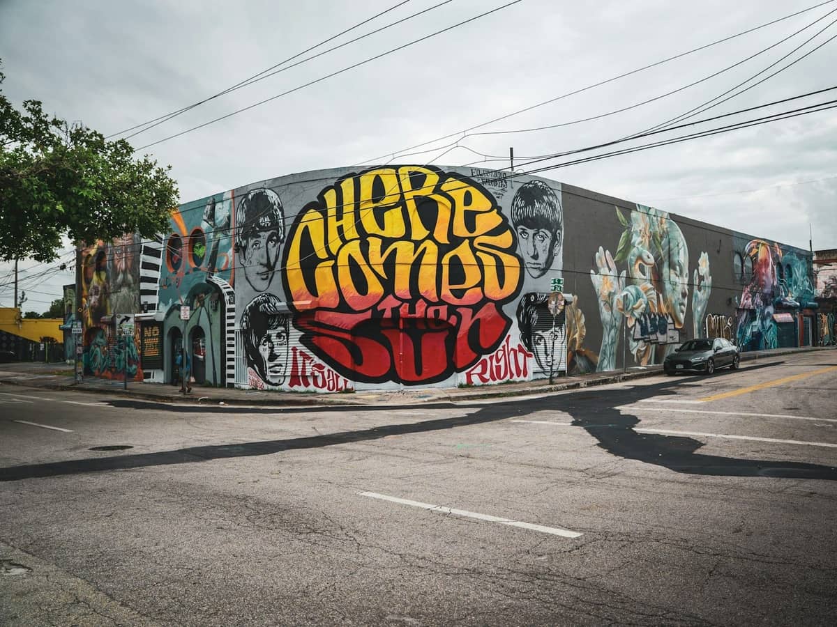 Colorful mural-covered warehouse in Miami's Wynwood neighborhood, featuring large bold text 'Here Comes the Sun' and various painted portraits on the side of the building.