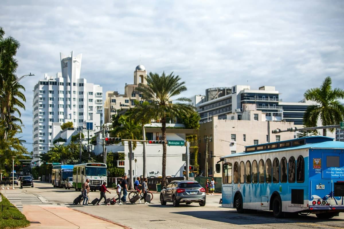 Street in Miami with palm trees, a city trolley bus, pedestrians, bikes, and Art Deco buildings under a partly cloudy sky.