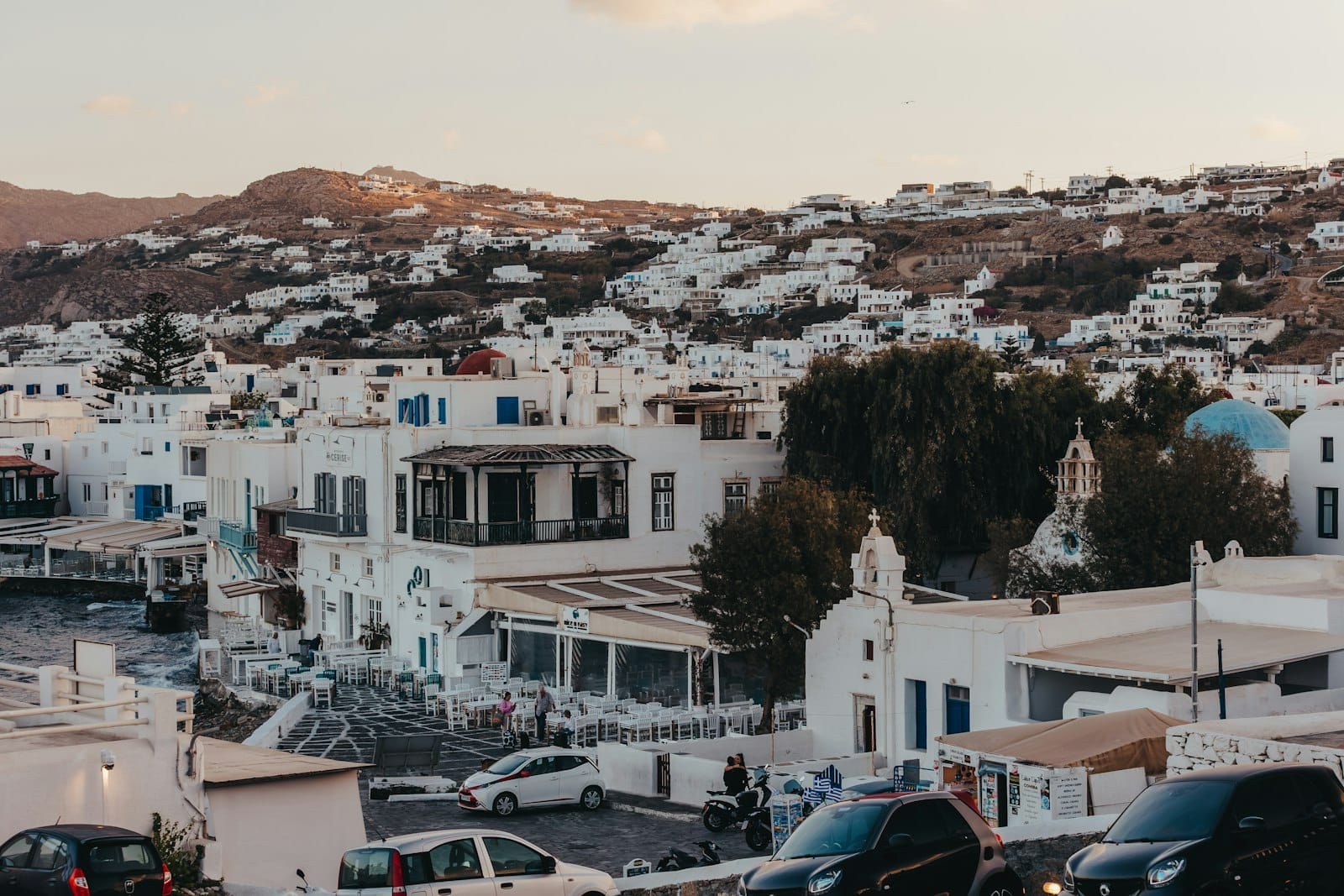 Vista del pueblo de Mykonos con coches aparcados, edificios encalados y colinas al fondo al atardecer