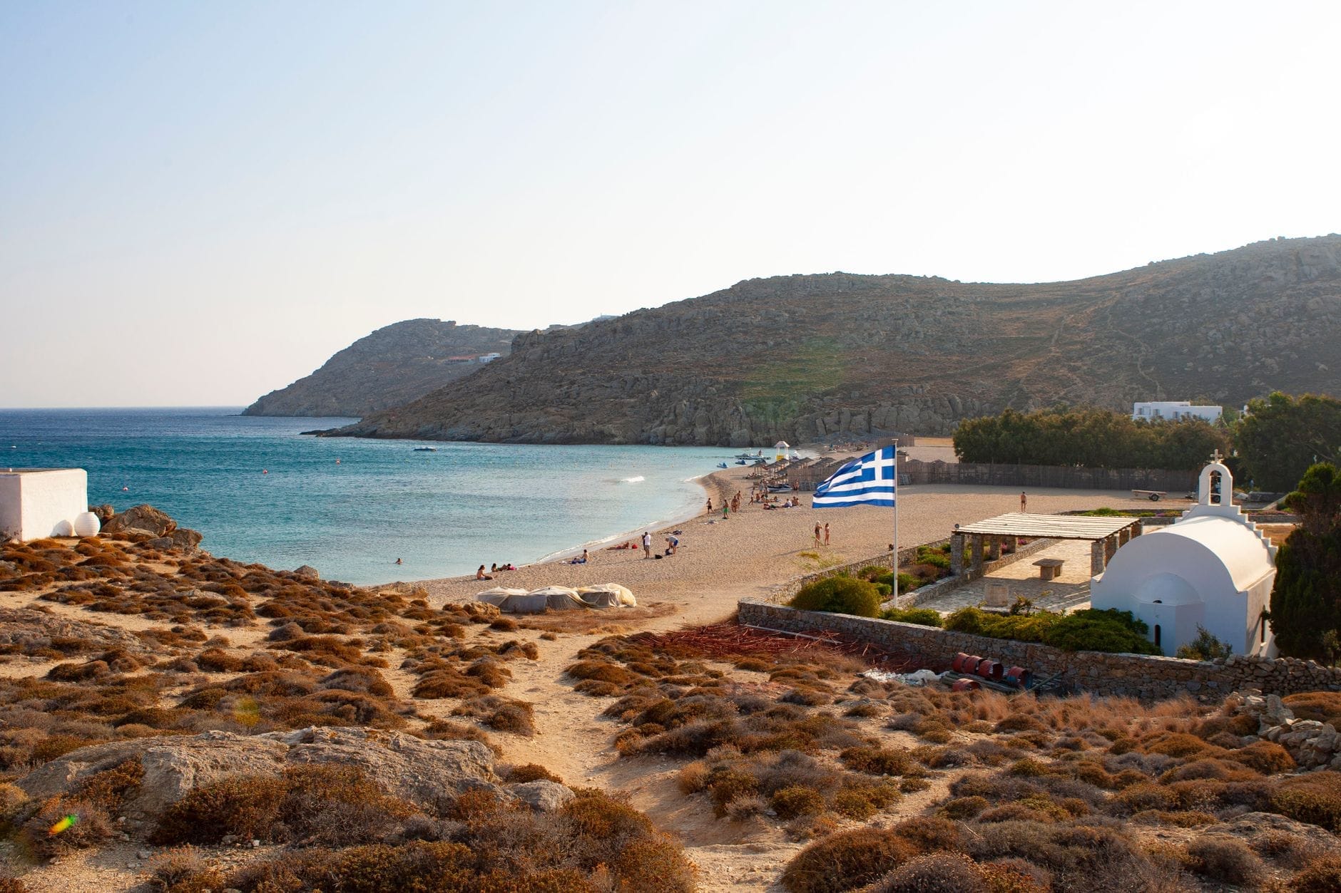Playa tranquila con pocas personas, bandera griega, capilla, edificios sencillos y colinas áridas con vistas a una bahía en Mykonos