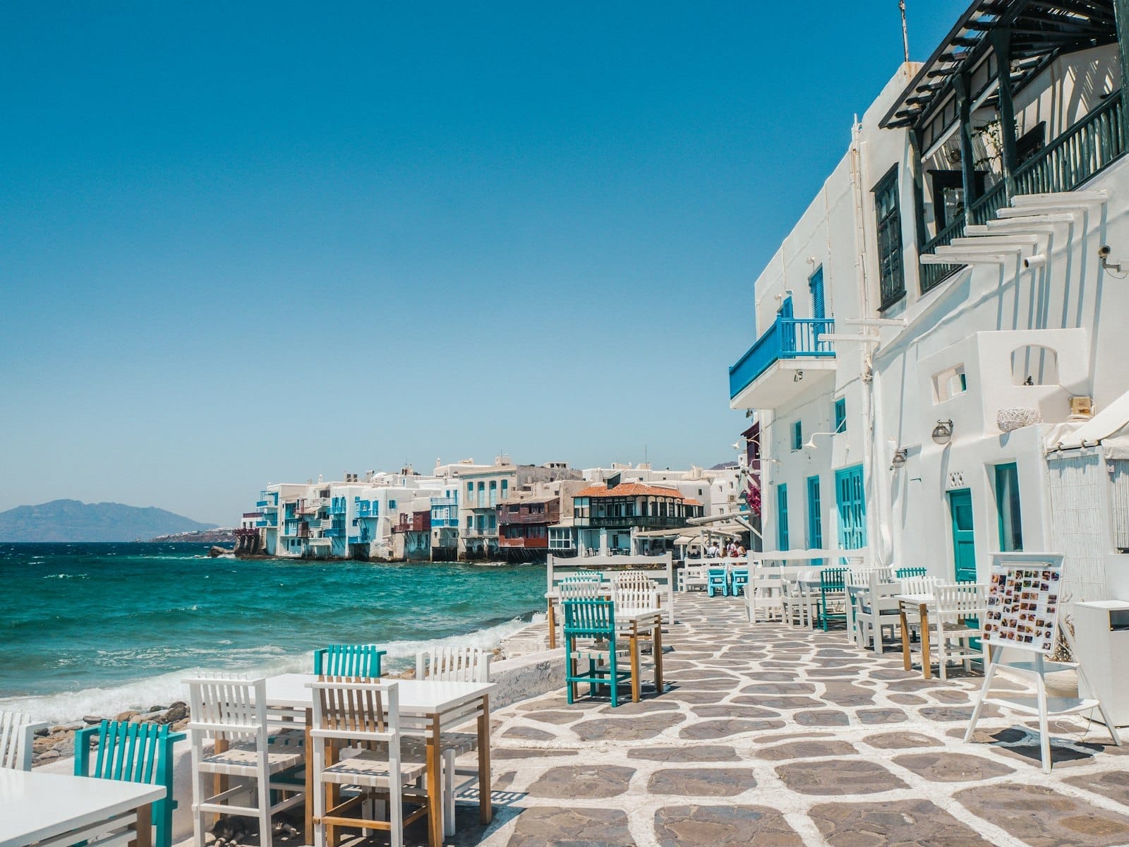 Vista costera de edificios blancos con detalles azules en Mykonos, con mesas y sillas al aire libre sobre un camino de piedra frente a aguas turquesas bajo un cielo azul despejado.
