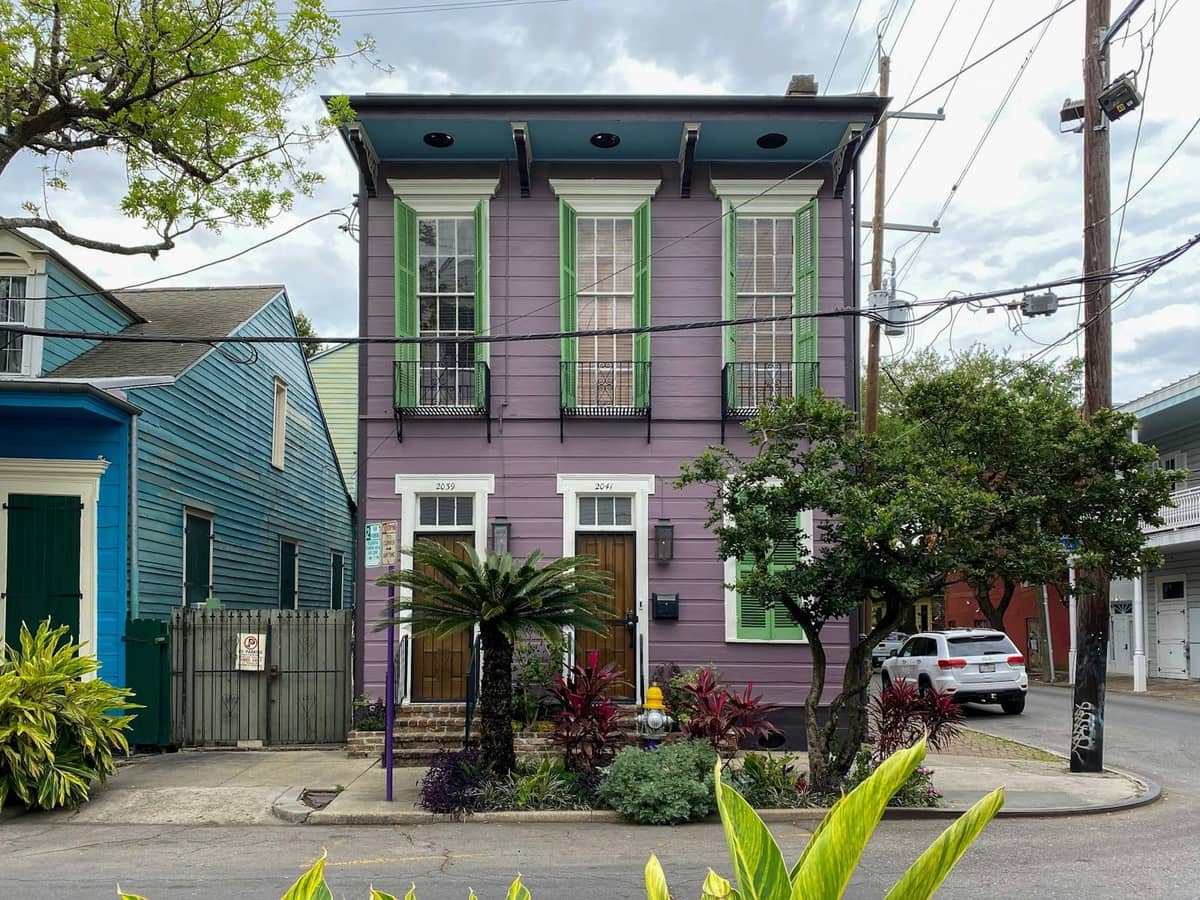 Una classica casa del Garden District di New Orleans dipinta di viola con finiture verdi, un rigoglioso giardino frontale e alte finestre su una strada alberata.