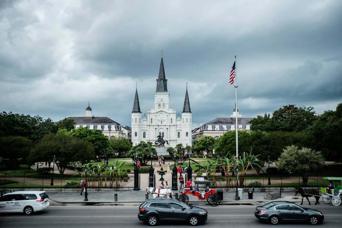 Ampia veduta di Jackson Square e della Cattedrale di St. Louis con le iconiche guglie e la statua equestre, auto e una carrozza a cavalli in primo piano.