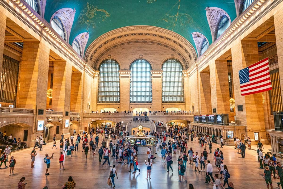 โถงหลักของ Grand Central Terminal ในย่าน Midtown Manhattan พร้อมเพดานรูปท้องฟ้าสีเขียวอมฟ้า ธงชาติอเมริกัน หน้าต่างโค้งขนาดใหญ่ และผู้คนมากมาย