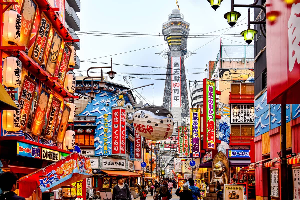 Daytime view of Shinsekai with Tsutenkaku Tower, colorful shop signs, lanterns, and people strolling the lively Osaka street.