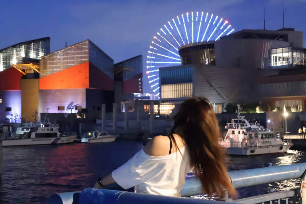 View of Osaka Aquarium Kaiyukan's colorful exterior and illuminated Tempozan Ferris Wheel at night, seen from the harbor with boats in the foreground.