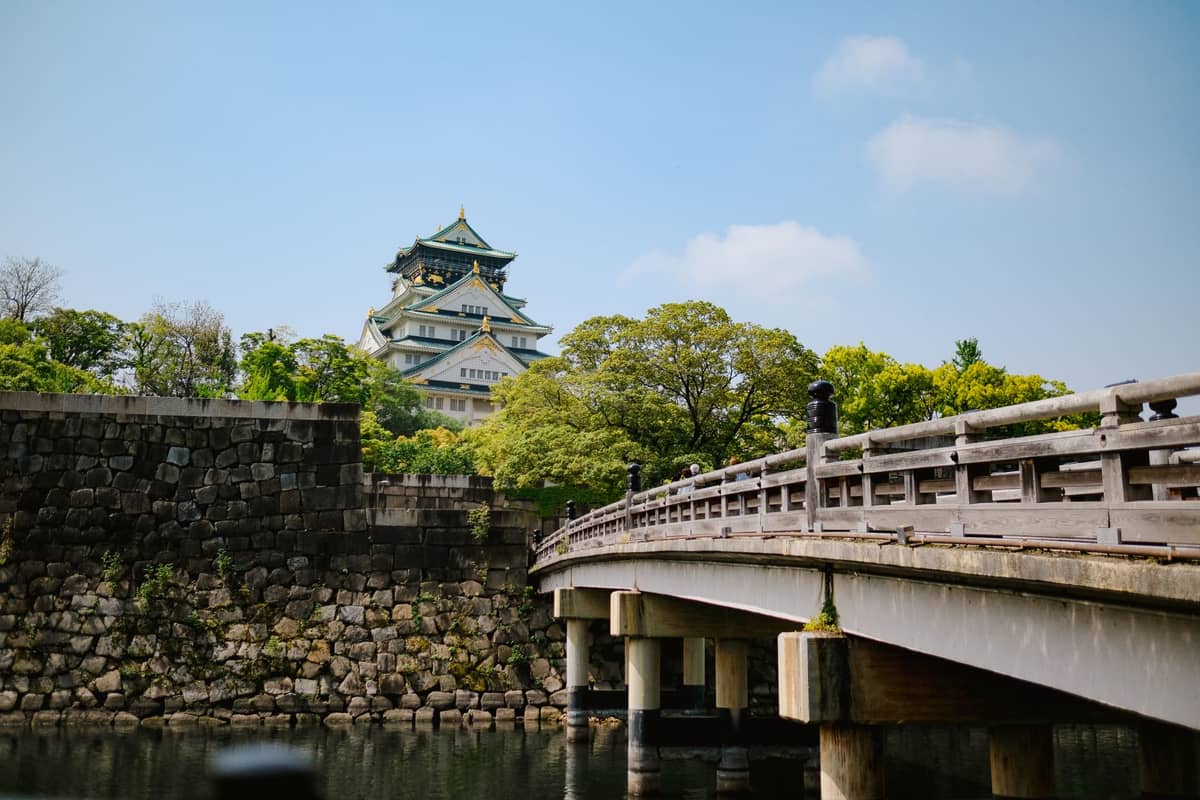 Osaka Castle in the background with a wooden bridge leading toward it, surrounded by greenery and a bright blue sky.