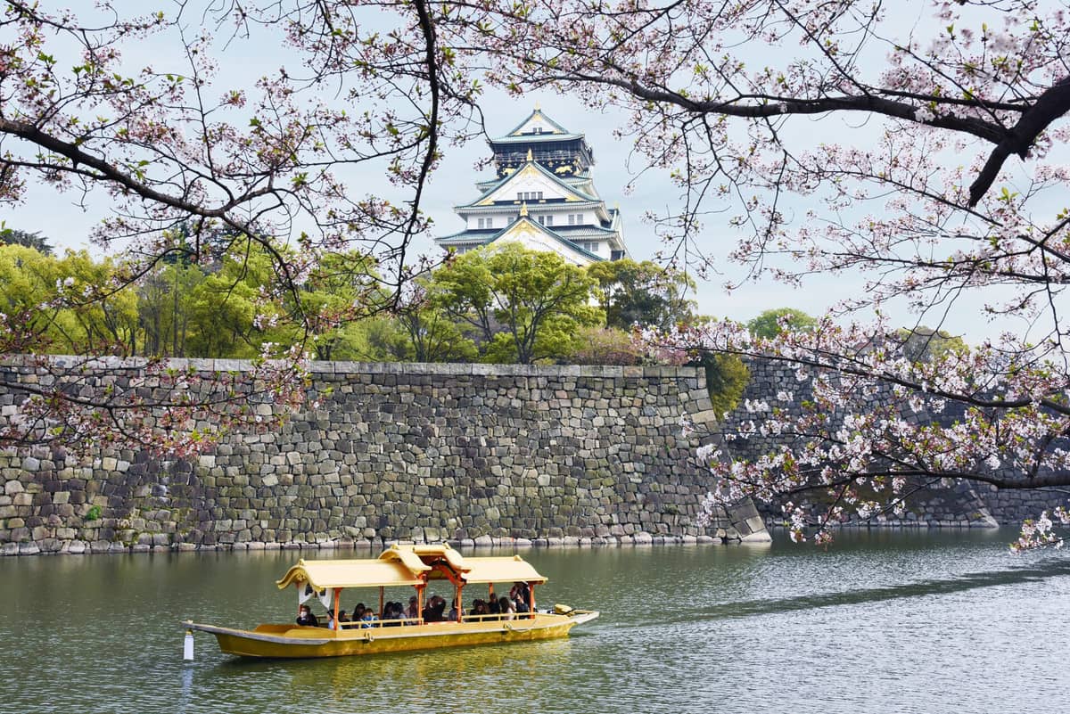 Osaka Castle framed by cherry blossoms, with a yellow sightseeing boat on the moat and stone walls clearly visible.