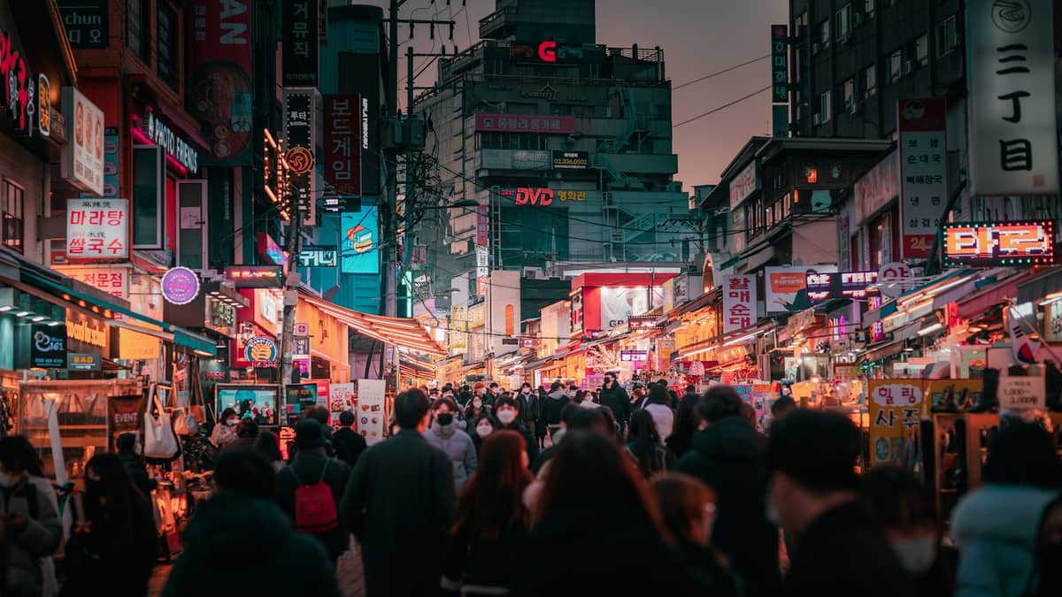 Movimentada cena de rua em Seul ao entardecer em um distrito mais moderno, com luzes de néon, letreiros vibrantes e multidões explorando lojas e restaurantes.