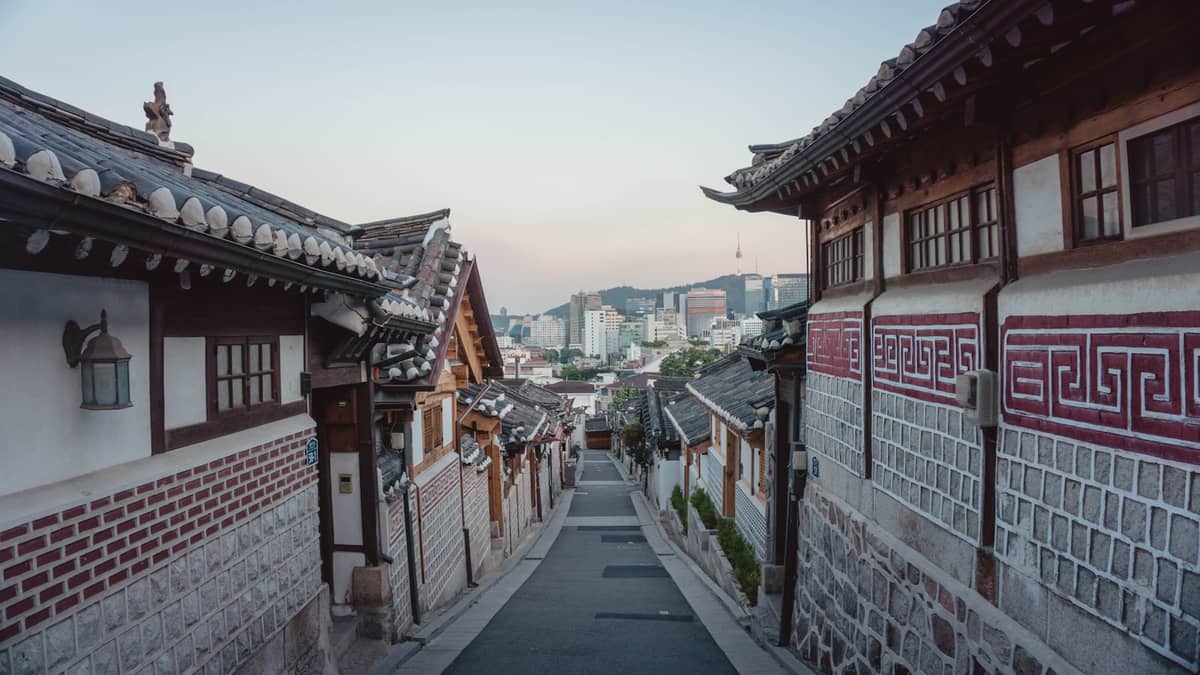 Rua tranquila ladeada por casas hanok tradicionais na Vila Hanok de Bukchon, em Seul, com o skyline da cidade e a Torre Namsan ao fundo.