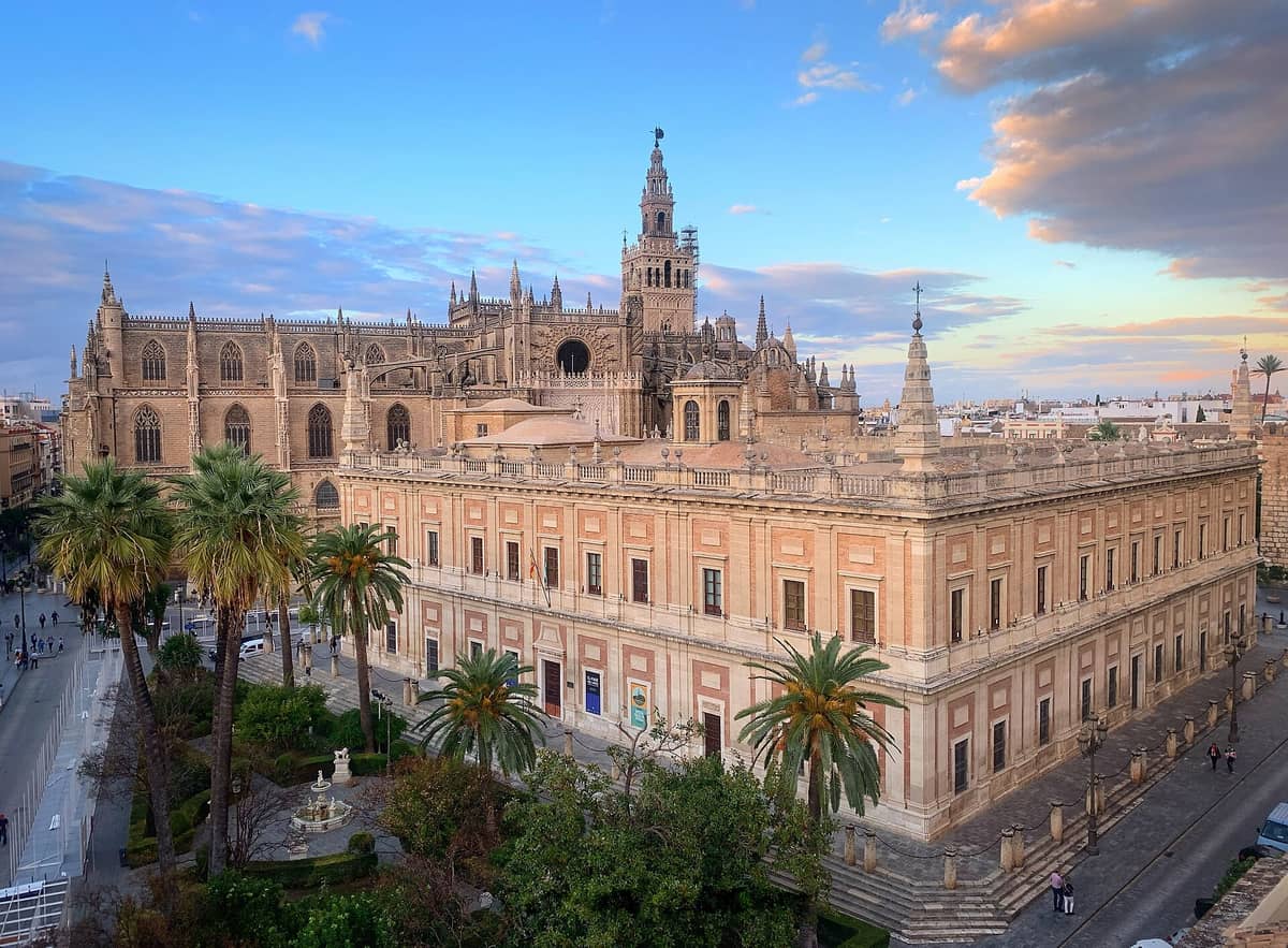 วิวทิวทัศน์ของมหาวิหาร Seville พร้อมหอคอย La Giralda ต้นปาล์ม และ Archivo de Indias ในยามพระอาทิตย์ตก