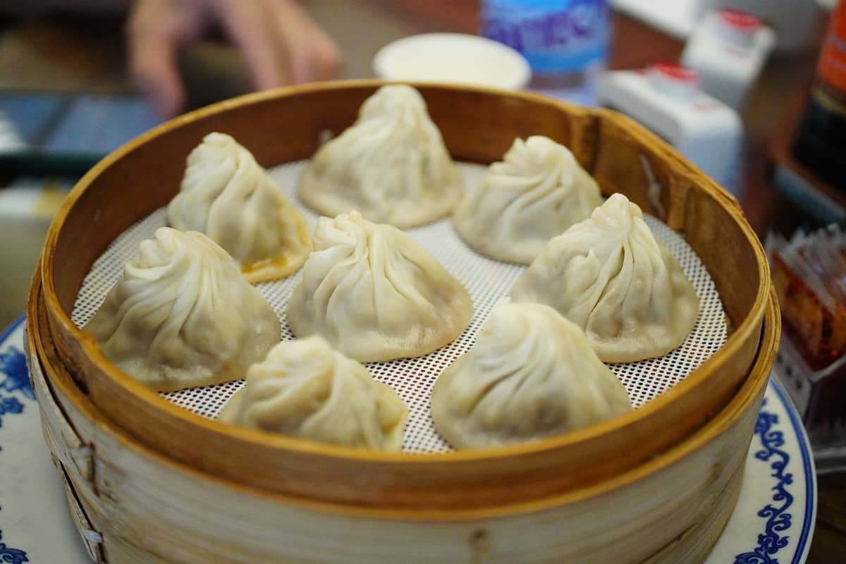 A bamboo steamer basket filled with classic Shanghai soup dumplings (xiaolongbao) on a table in a bright restaurant setting.