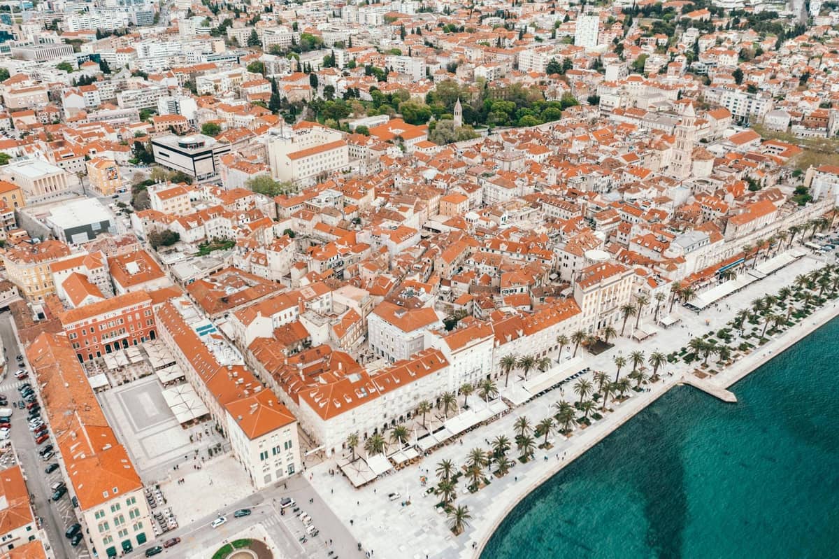 Luftaufnahme der Altstadt von Split mit Terrakotta-Dächern, historischen Gebäuden, palmengesäumter Uferpromenade und dem klaren blauen Adriatischen Meer neben der Promenade.