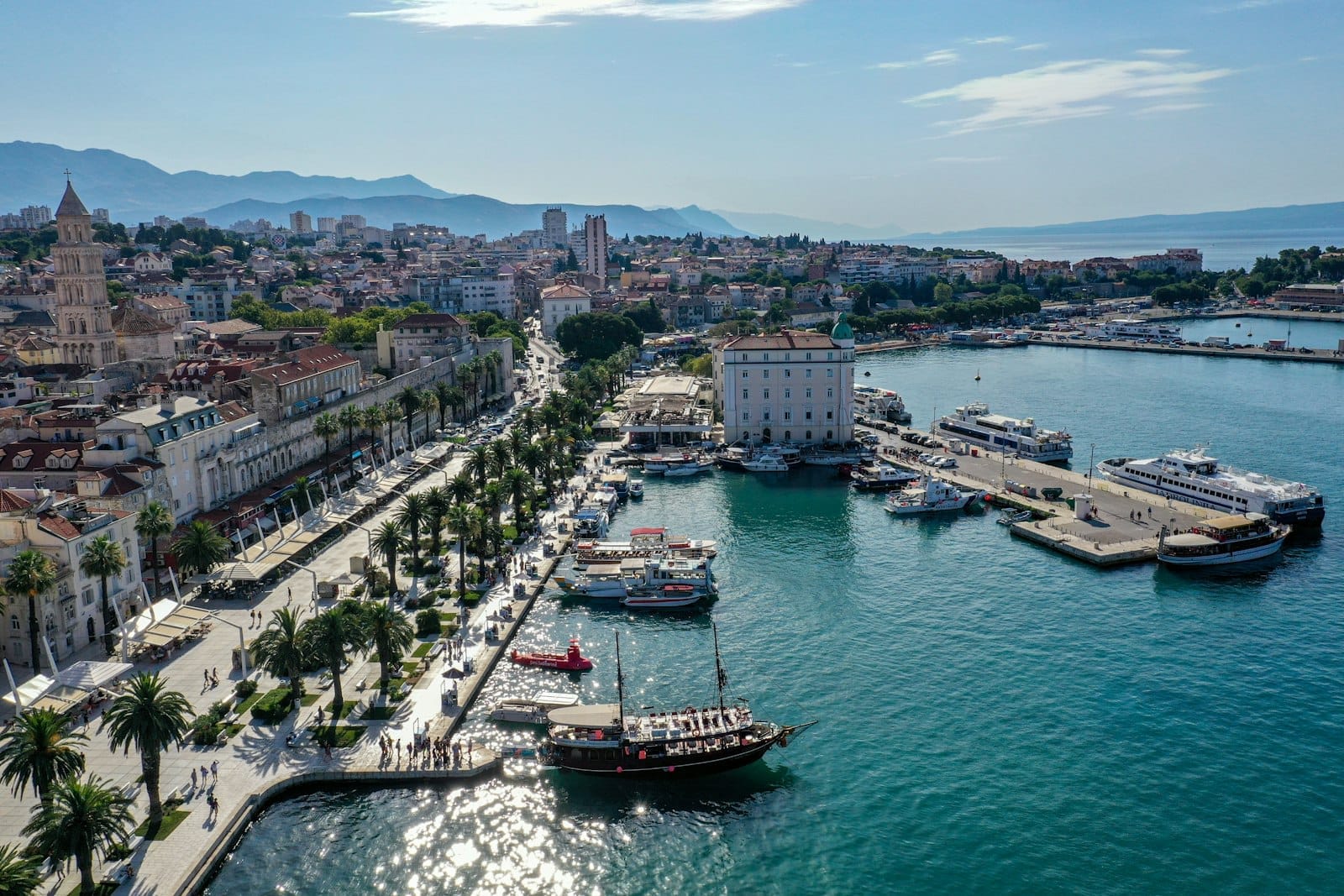 Vista aérea panorâmica do porto de Split com barcos, o calçadão à beira-mar ladeado de palmeiras, prédios históricos e morros ao fundo em um dia ensolarado.