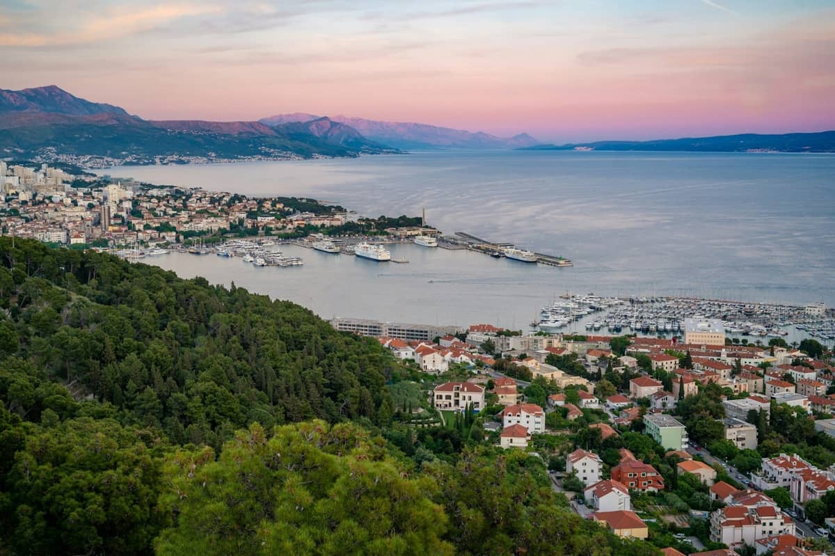 Panoramablick auf die Küste von Split mit dem Marjan-Hügel, Stadtvierteln, Marina und fernen Bergen bei Sonnenuntergang.
