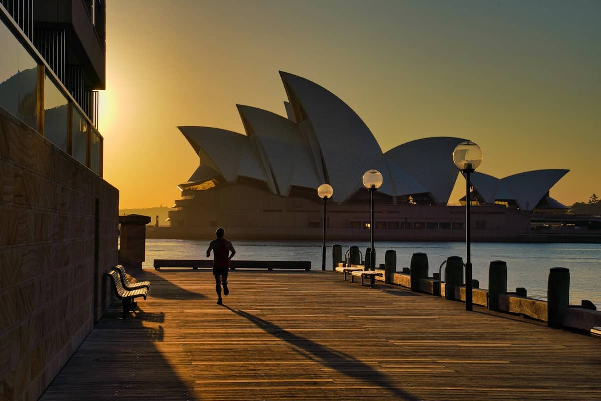 Sydney Opera House im Morgenlicht mit einem Jogger auf der östlichen Promenade und leeren Bänken, von Circular Quay aus gesehen