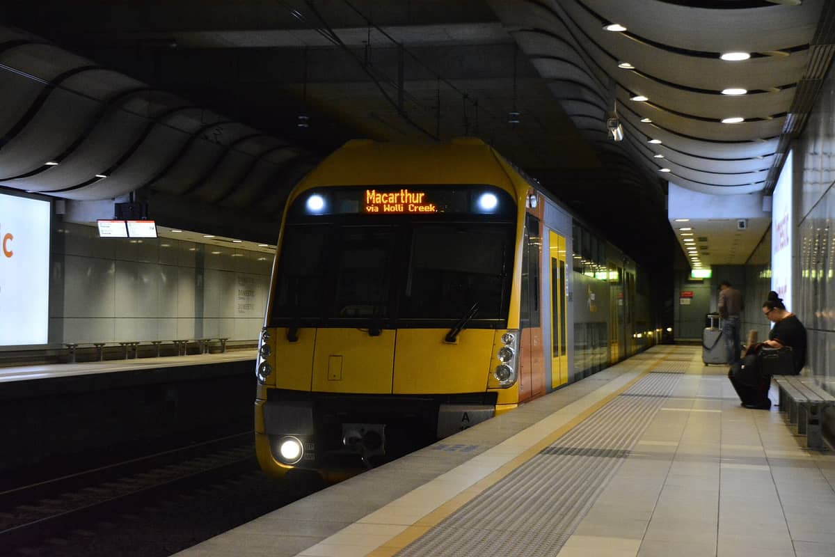 Ein Sydney Trains-Zug am Bahnsteig der Station Domestic Airport, mit Fahrgästen und Gepäck in einem modernen unterirdischen Terminal.