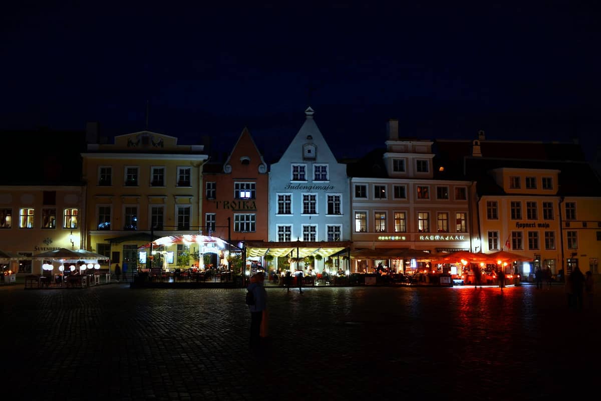 Cafés y restaurantes al aire libre iluminados por la noche en la plaza del casco histórico de Tallinn, con coloridos edificios medievales al fondo.
