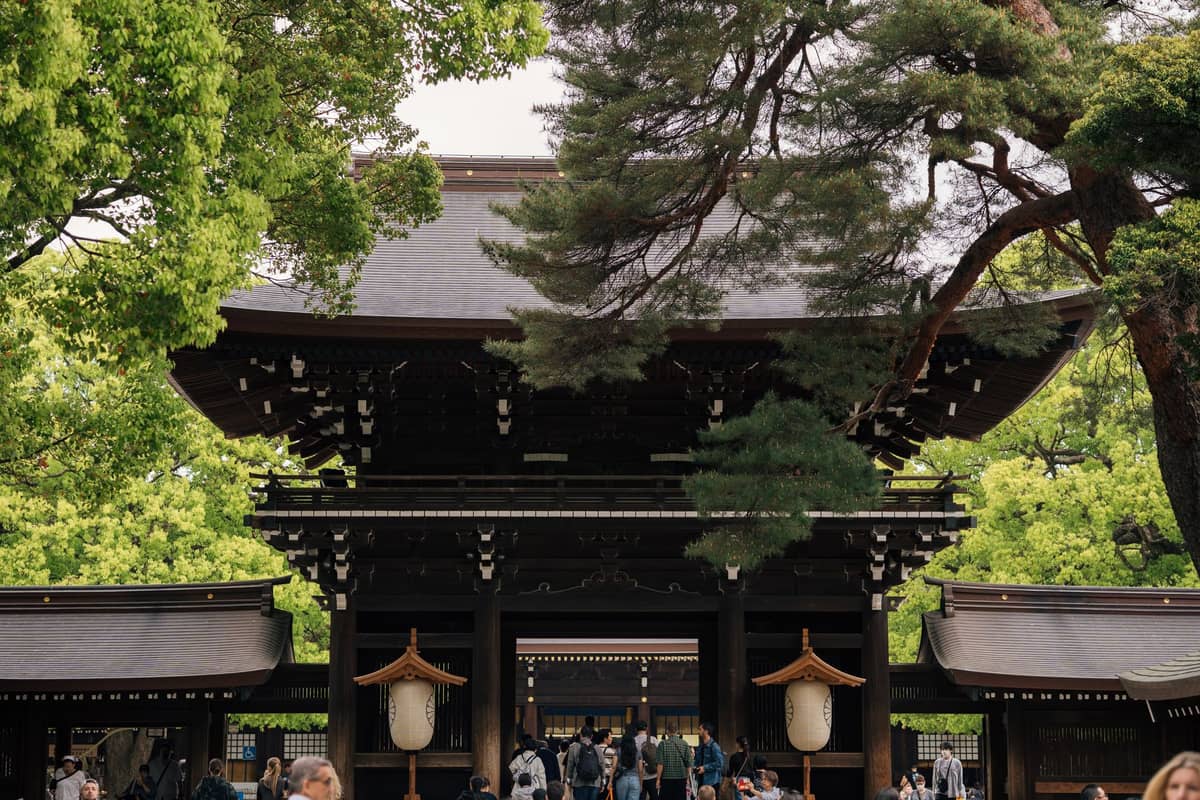 The entrance to Meiji Jingu Shrine in Tokyo with a large wooden gate, trees, and visitors walking beneath the gateway.