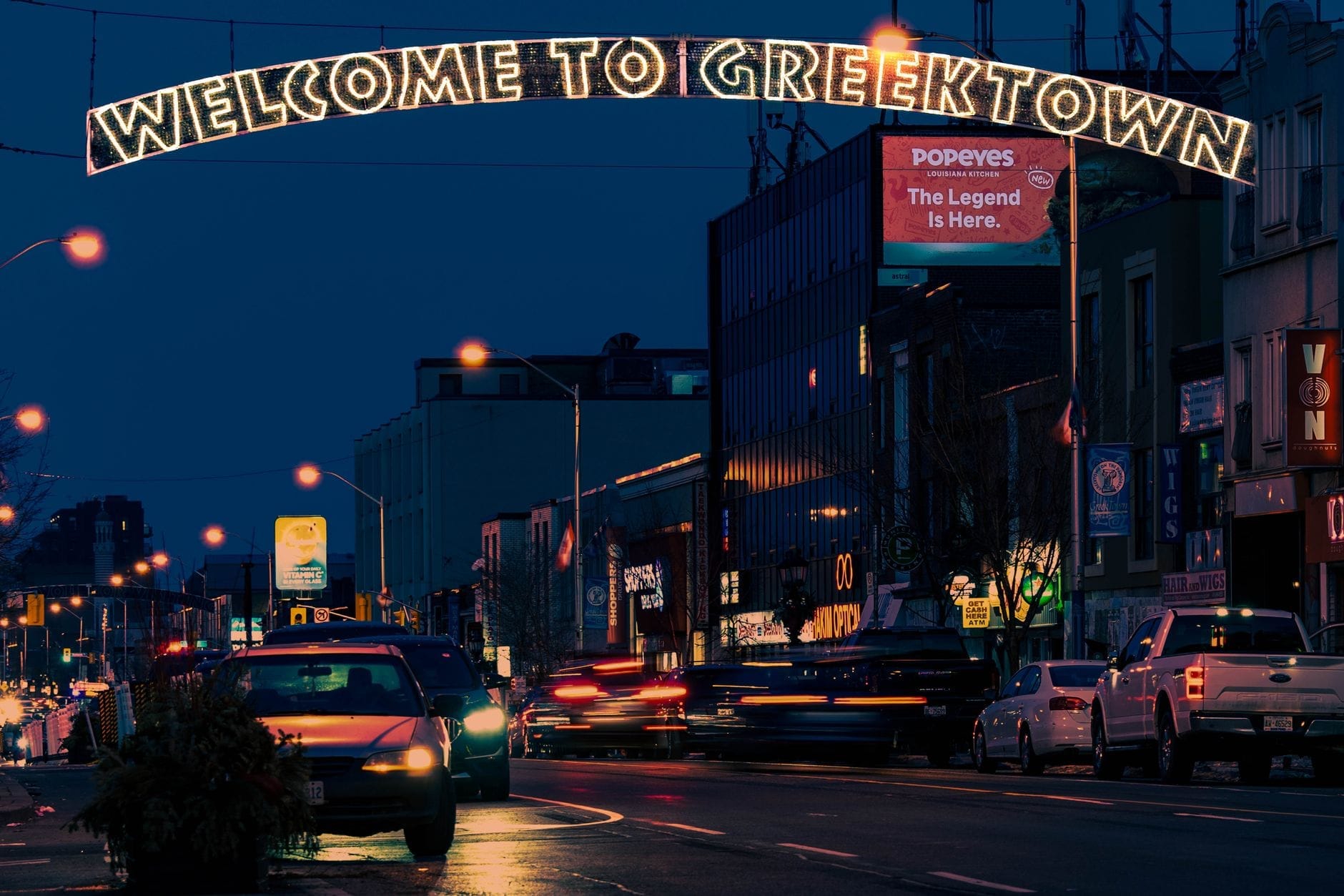 Cena noturna de rua em Greektown, Toronto, com um letreiro luminoso 'Welcome to Greektown' e carros na estrada.