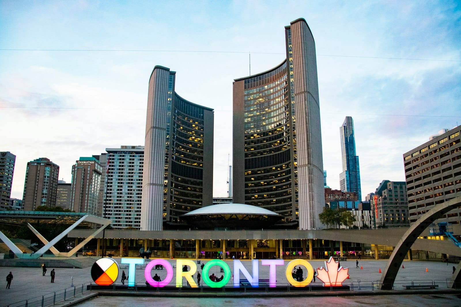 A Câmara Municipal de Toronto e o colorido letreiro iluminado de Toronto em Nathan Phillips Square, com os edifícios da cidade ao fundo ao entardecer.