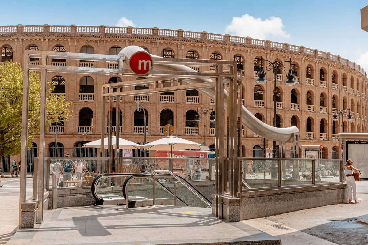 Straßenebenener Eingang zur Metro Valencia mit großem rotem Metro-Schild und historischem Stierkampfarena-Gebäude im Hintergrund an einem sonnigen Tag