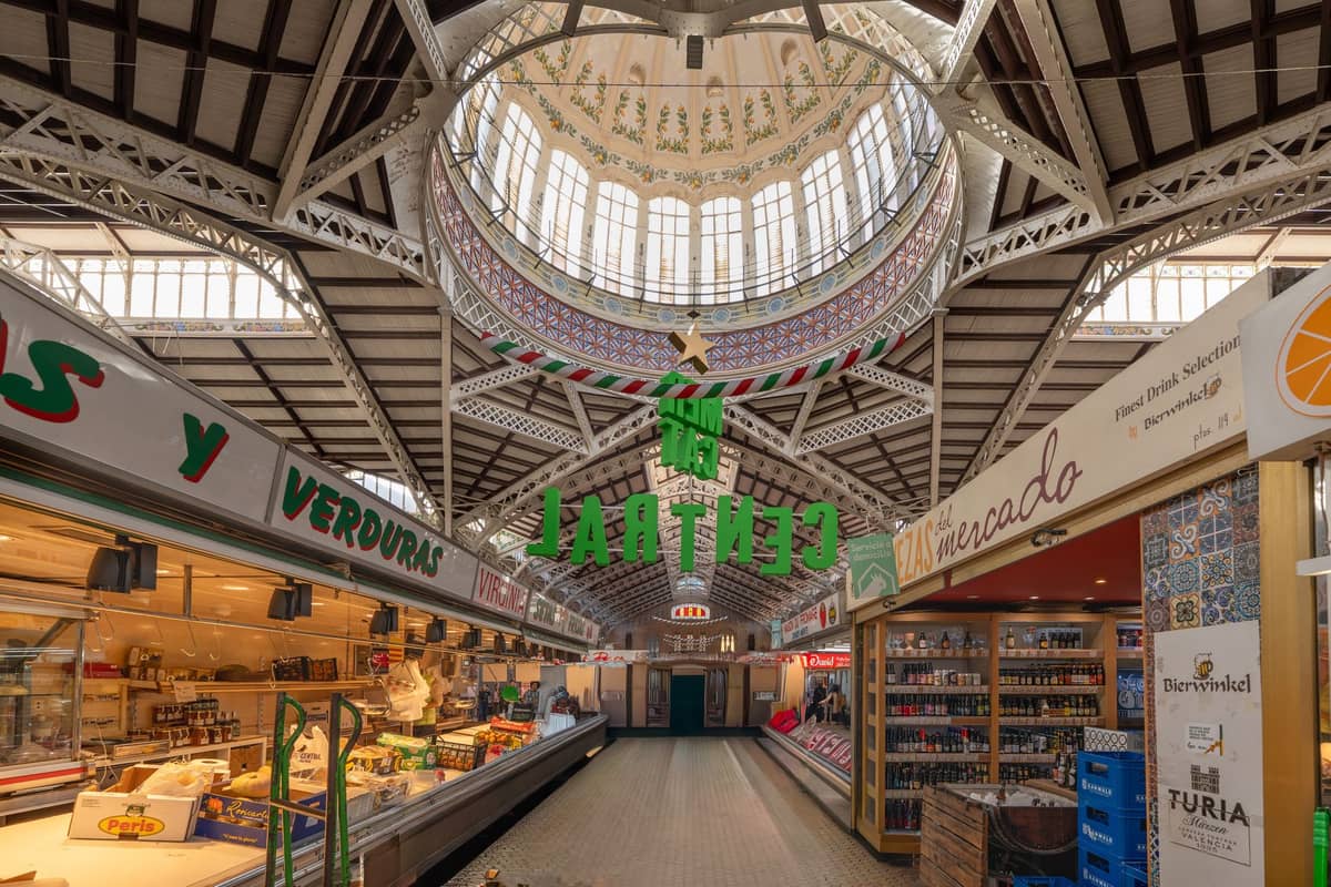 Weitwinkelansicht des Innenraums des Mercado Central de Valencia mit Marktständen, den verzierten Buntglasfenstern und der markanten modernistischen Zentralkuppel.