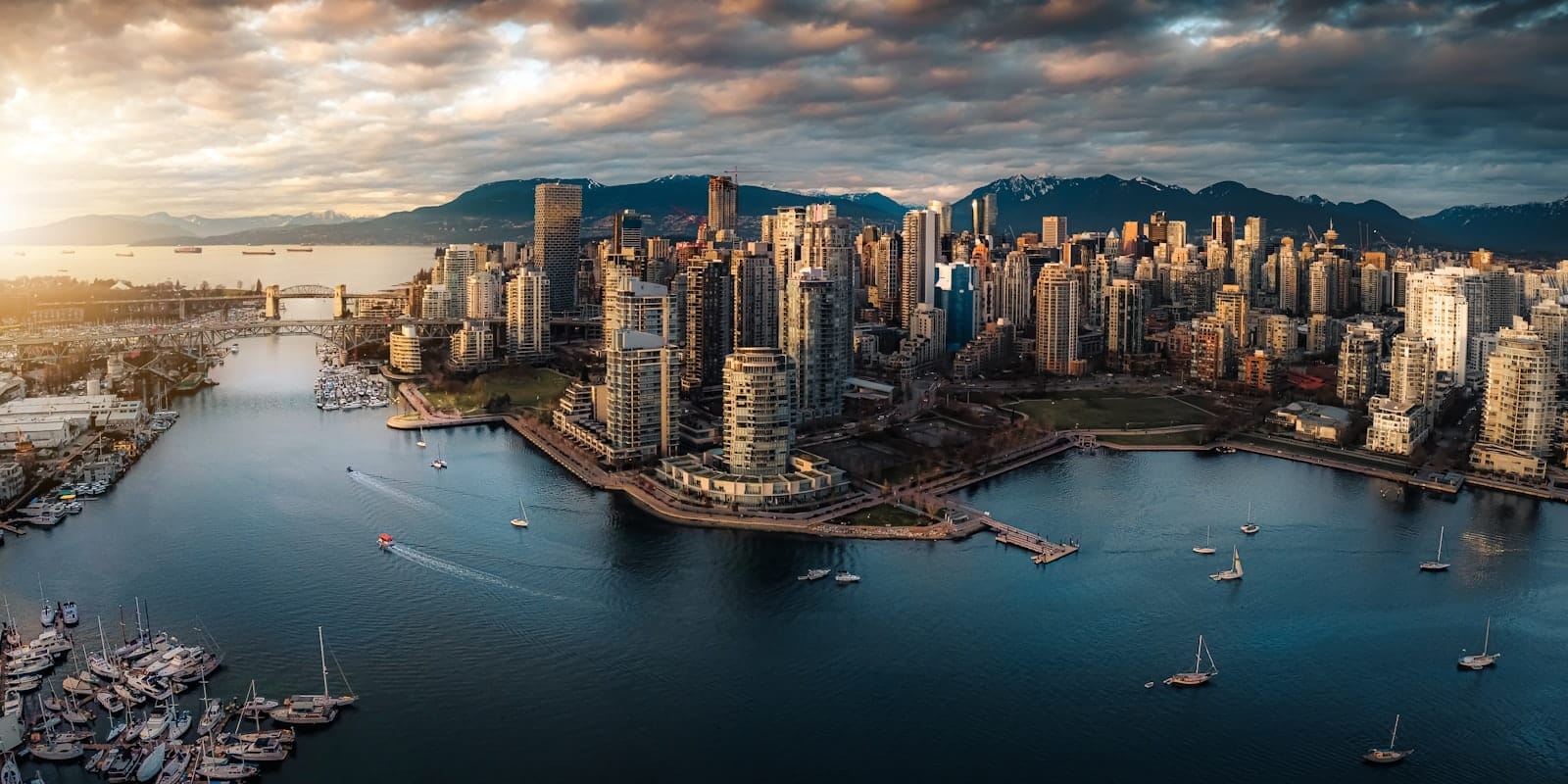 Panorama aéreo do centro de Vancouver ao pôr do sol com orla, marina, skyline e montanhas ao fundo sob nuvens dramáticas.