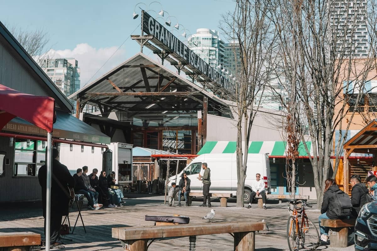 วิวภายนอกทางเข้า Granville Island Public Market พร้อมผู้คนนั่งและเดิน รถตู้สีขาว และอาคารในเมืองเป็นฉากหลัง
