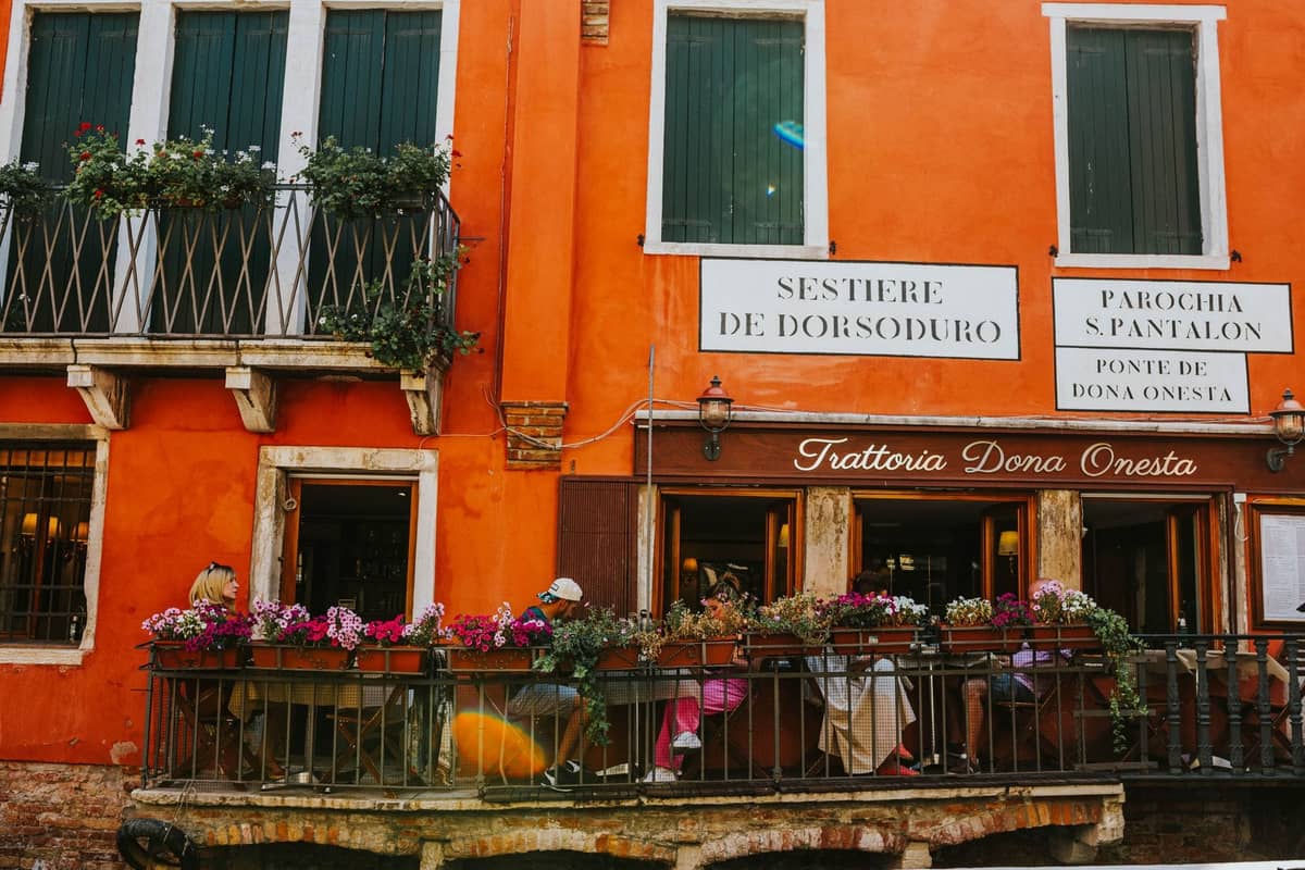 Leuchtend orangefarbenes Gebäude in Venedig mit Schildern für Sestiere de Dorsoduro und Trattoria Dona Onesta, Menschen beim Essen auf der Terrasse
