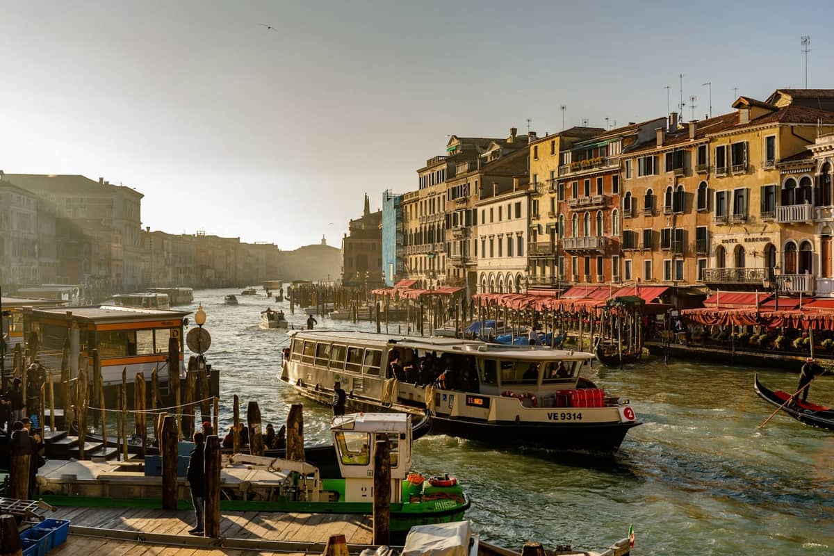 Venedigs Canal Grande mit Vaporetto-Wasserbus, vertäuten Booten, historischen bunten Gebäuden und lebhafter Uferpromenade unter klarem Himmel.