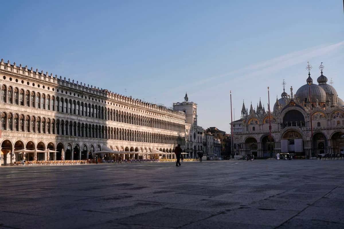 Weite Aussicht auf eine leere Piazza San Marco bei Sonnenaufgang, mit den Procuratie und der Basilika San Marco unter klarem Himmel.