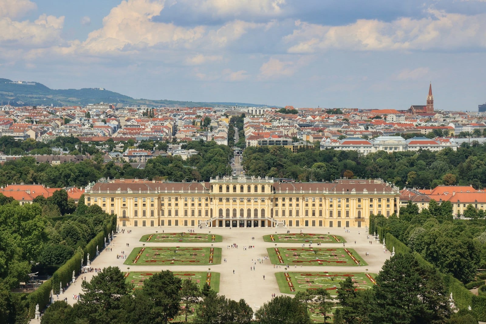 Vista aérea del Palacio de Schönbrunn con sus jardines, avenidas arboladas y el paisaje urbano de Viena al fondo bajo un cielo azul.