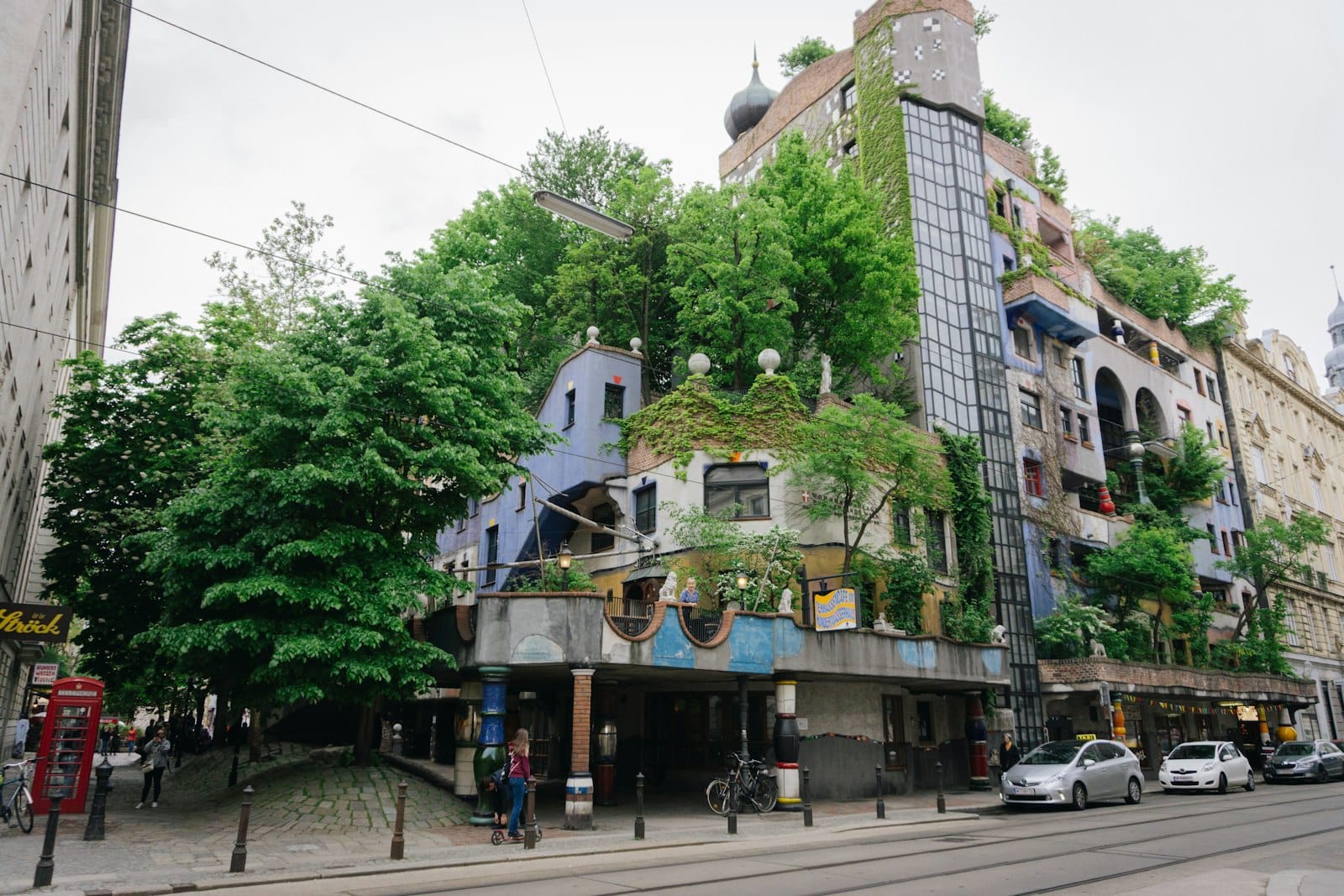 Vista amplia de la colorida Hundertwasserhaus de Viena, con vegetación en el edificio y personas caminando cerca.