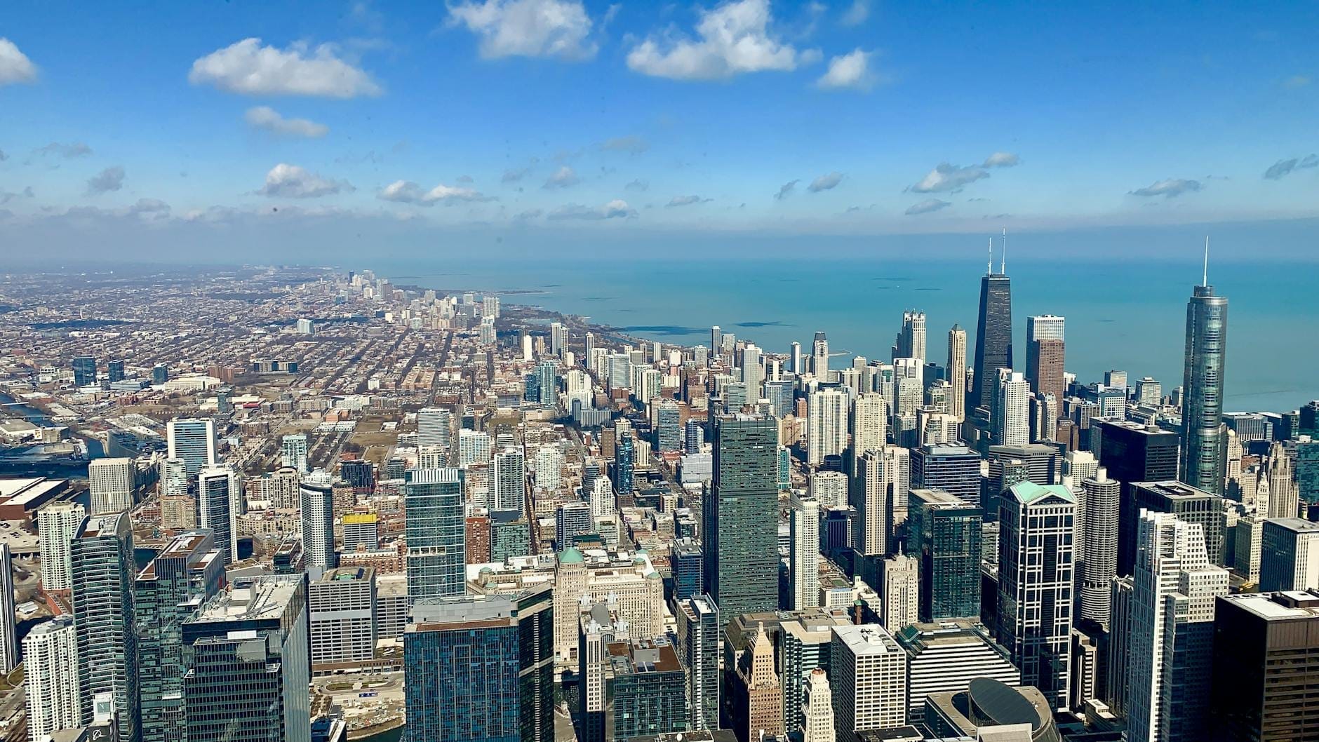 Vista panoramica dello skyline di Chicago e del Lago Michigan in una giornata di sole, ripresa da un'alta terrazza panoramica con cielo blu e nuvole sparse.