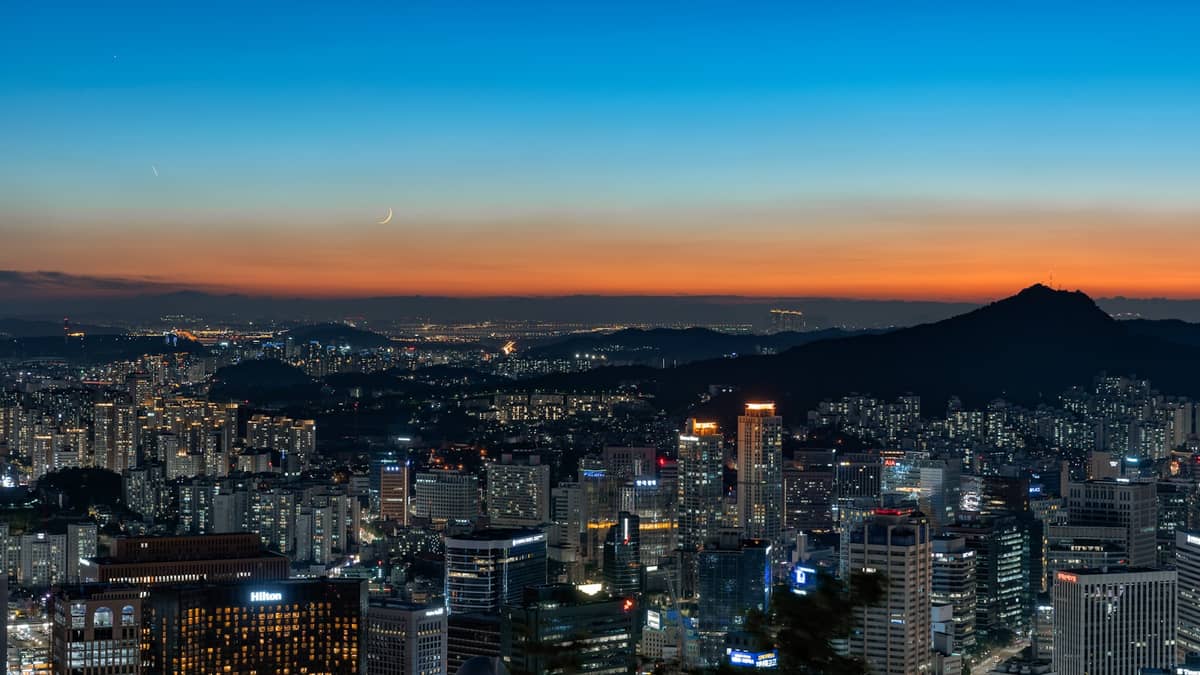 Panorâmica noturna de Seul com arranha-céus, montanhas ao fundo e um céu colorido ao pôr do sol, transmitindo a atmosfera moderna e expansiva da cidade.