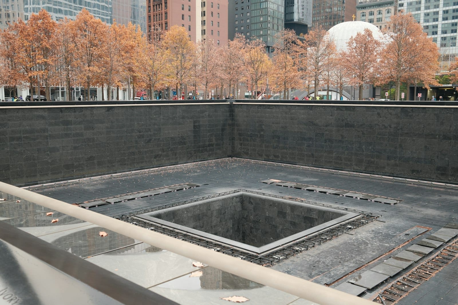 La vasca riflettente del National September 11 Memorial con alberi autunnali e palazzi della città sullo sfondo in una giornata nuvolosa.