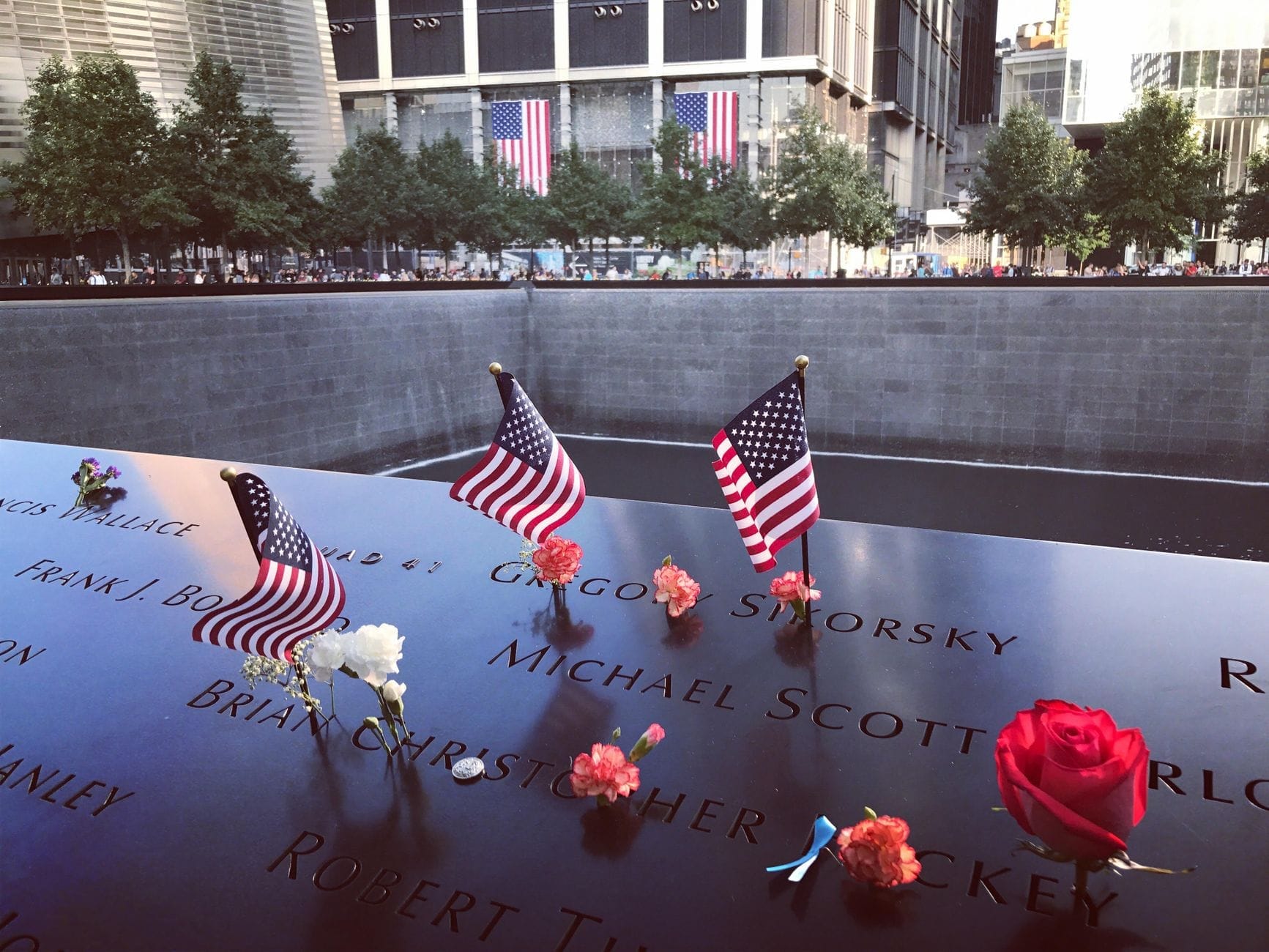 Alcune piccole bandiere americane e fiori posati sulle iscrizioni dei nomi al bordo del Memoriale dell'11 Settembre, con le vasche e gli edifici della città sullo sfondo.