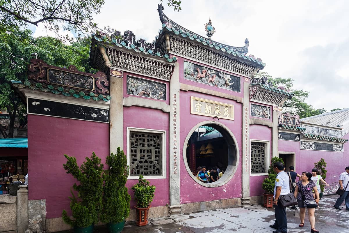 Frontansicht des Eingangs des A-Ma-Tempels in Macau mit rosa Wänden, kunstvollen Schnitzereien und Besuchern, die tagsüber draußen spazieren.