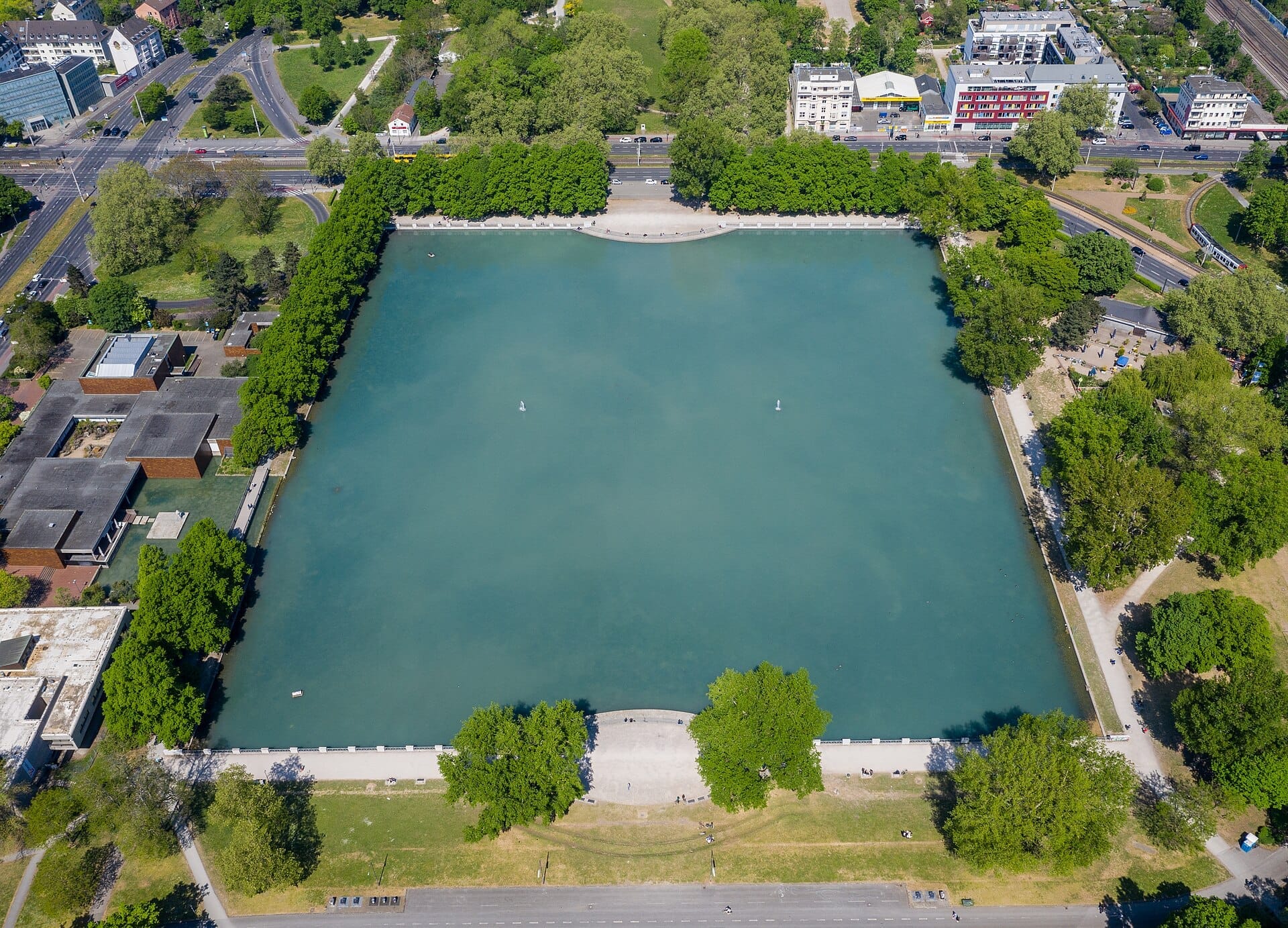 มุมมองทางอากาศของ Aachener Weiher แสดงให้เห็นสระน้ำเทียมขนาดใหญ่ที่ล้อมรอบด้วยต้นไม้สีเขียว เส้นทางเดินเท้า และอาคารใกล้เคียงในเมือง Cologne