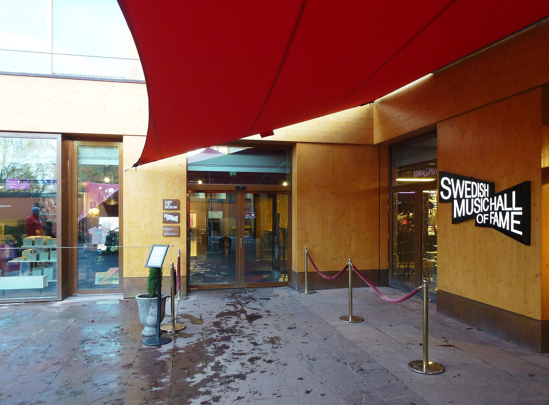 ทางเข้าด้านนอกของ ABBA The Museum และ Swedish Music Hall of Fame ป้ายชื่อชัด ประตูแก้ว แคนูปีสีแดงเหนือทางเดินหินในสตอกโฮล์ม