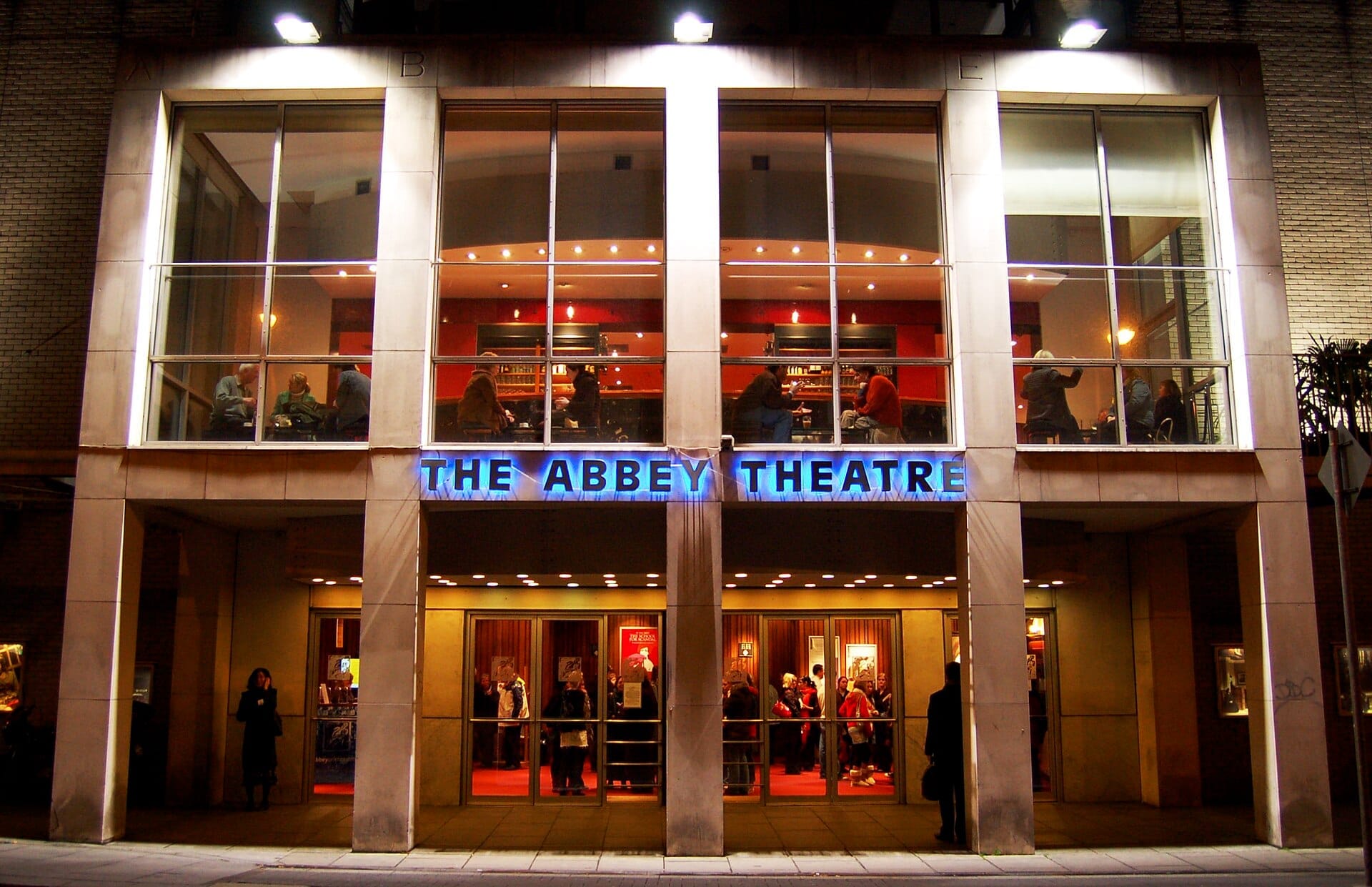 L'ingresso principale dell'Abbey Theatre a Dublino di notte, con insegne illuminate, grandi vetrate e persone visibili all'interno.